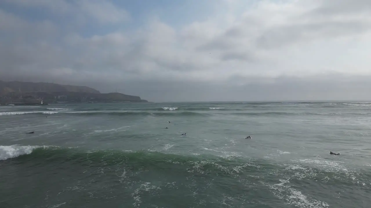 Wide aerial dolly shot of the calm sea in miraflores lima in peru with surfers on surfboards in the water and a view of the cloudy sky with mountains in the background