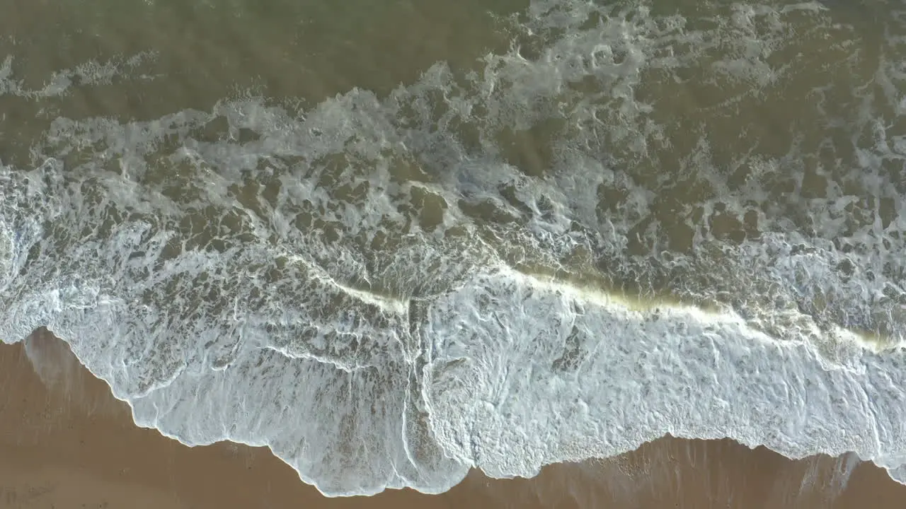 Aerial view of high powerful white foam waves slowly crashing on an empty sandy beach