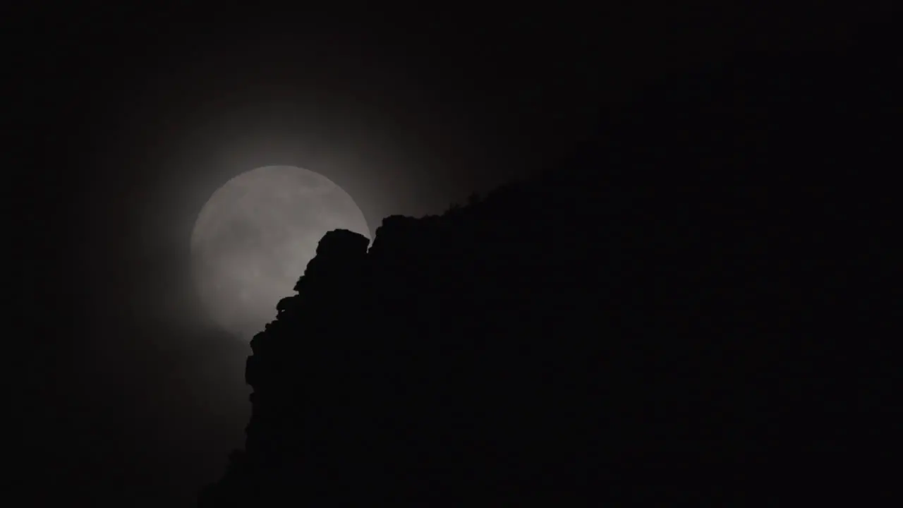 A full moon rises behind clouds and a rocky mountain foreground