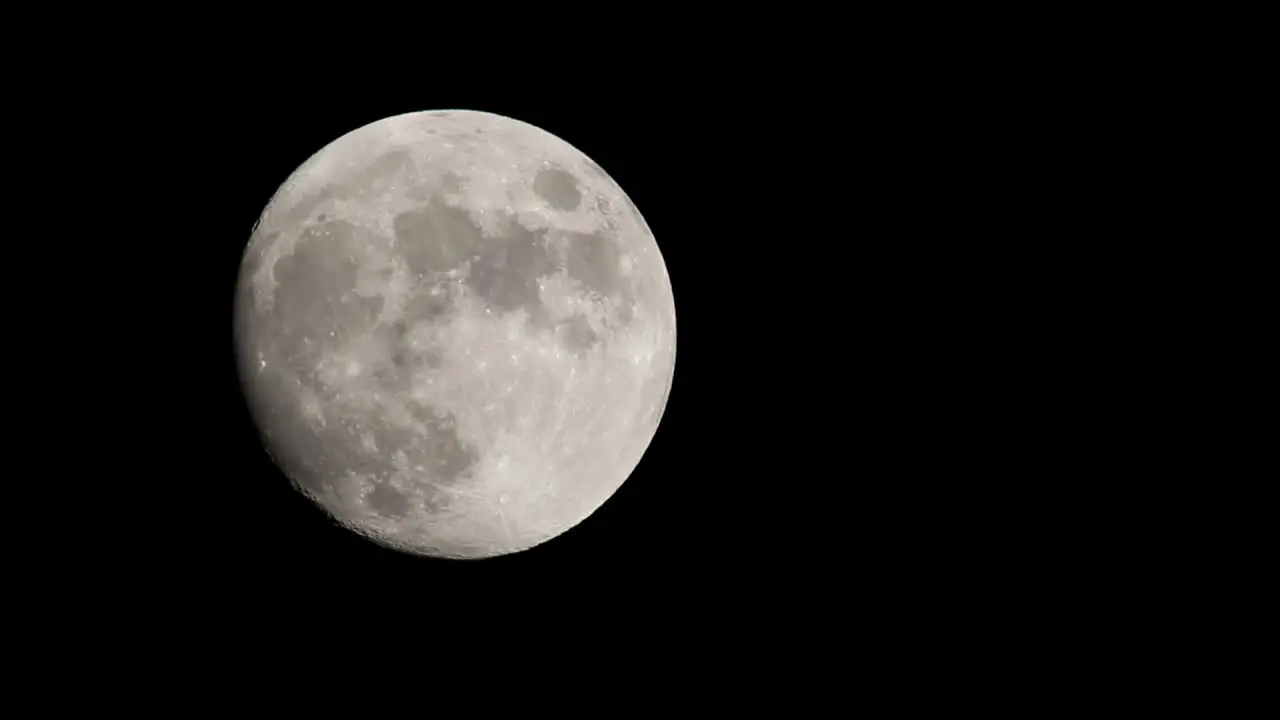 Bright full moon set against dark black sky moving from middle left of frame towards top center of frame