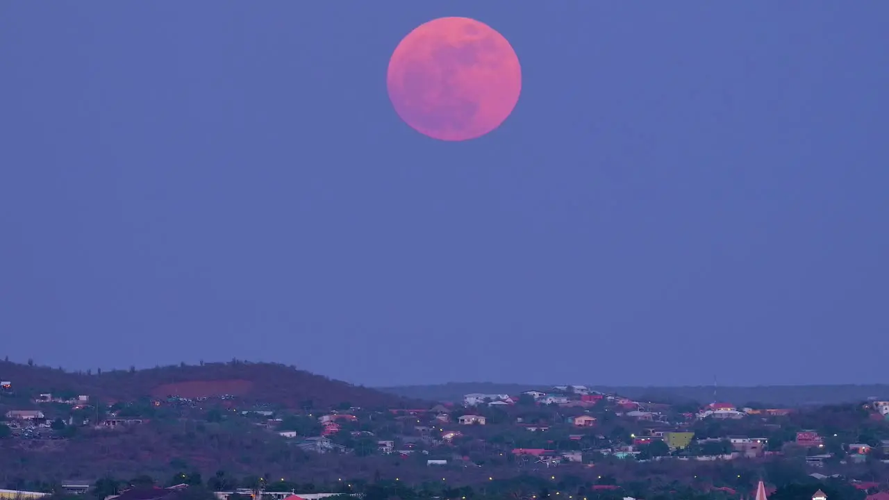 Pan up across homes on hillside of willemstad curacao near janwe church to supermoon pink and red