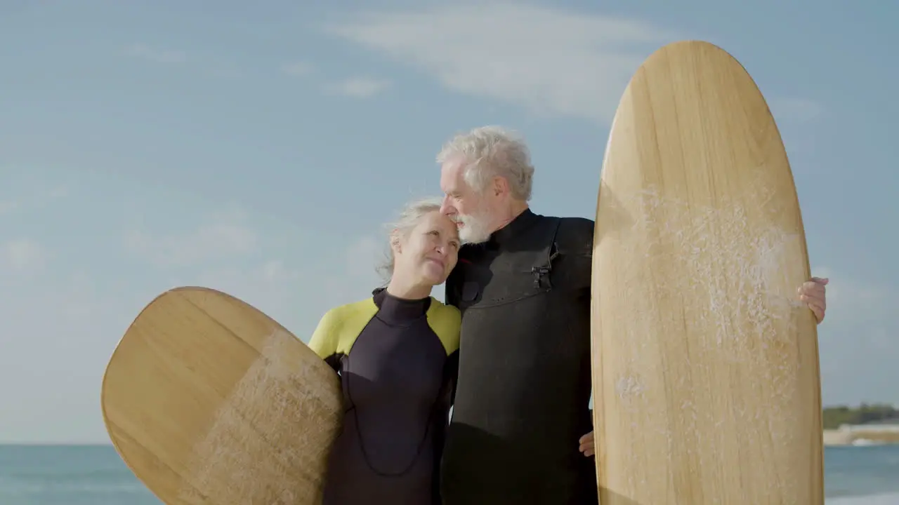 Front View Of A Senior Couple In Wetsuit With Surfboard Standing On The Sandy Beach Looking At The Camera And Kissing