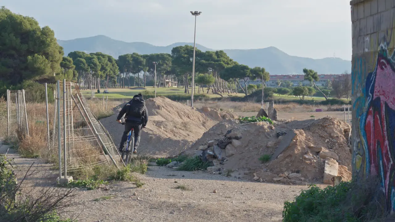 Cyclist cicles trough buildingsite around sand heaps  wire fence on one side and wall with graffitti on other side nice landscape with trees and mountains in distance