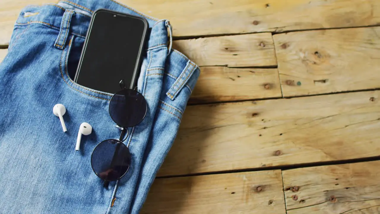 Close up of jeans with smartphone earphones and sunglasses on wooden background with copy space