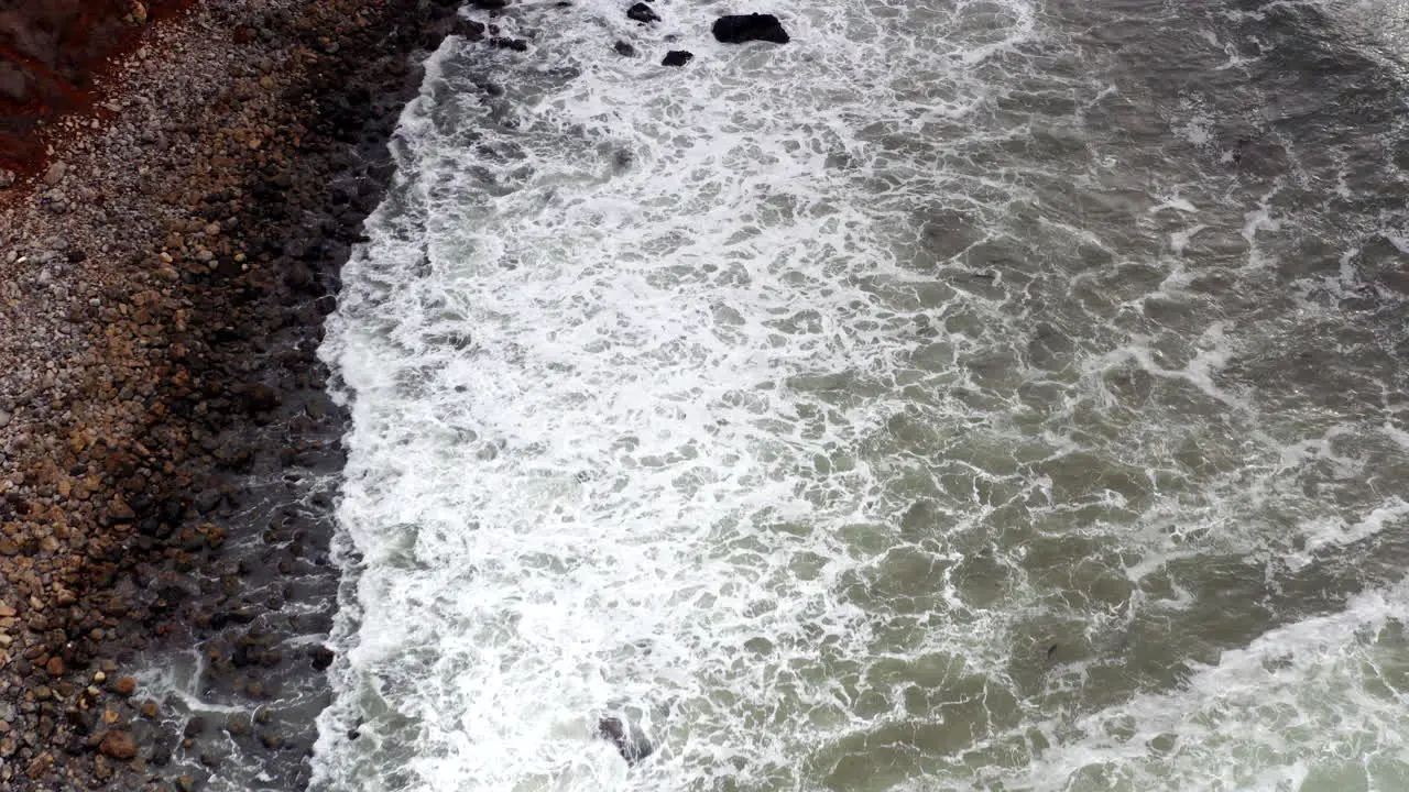 Aerial fly over of rocky beach with grey waves crashing onto shore