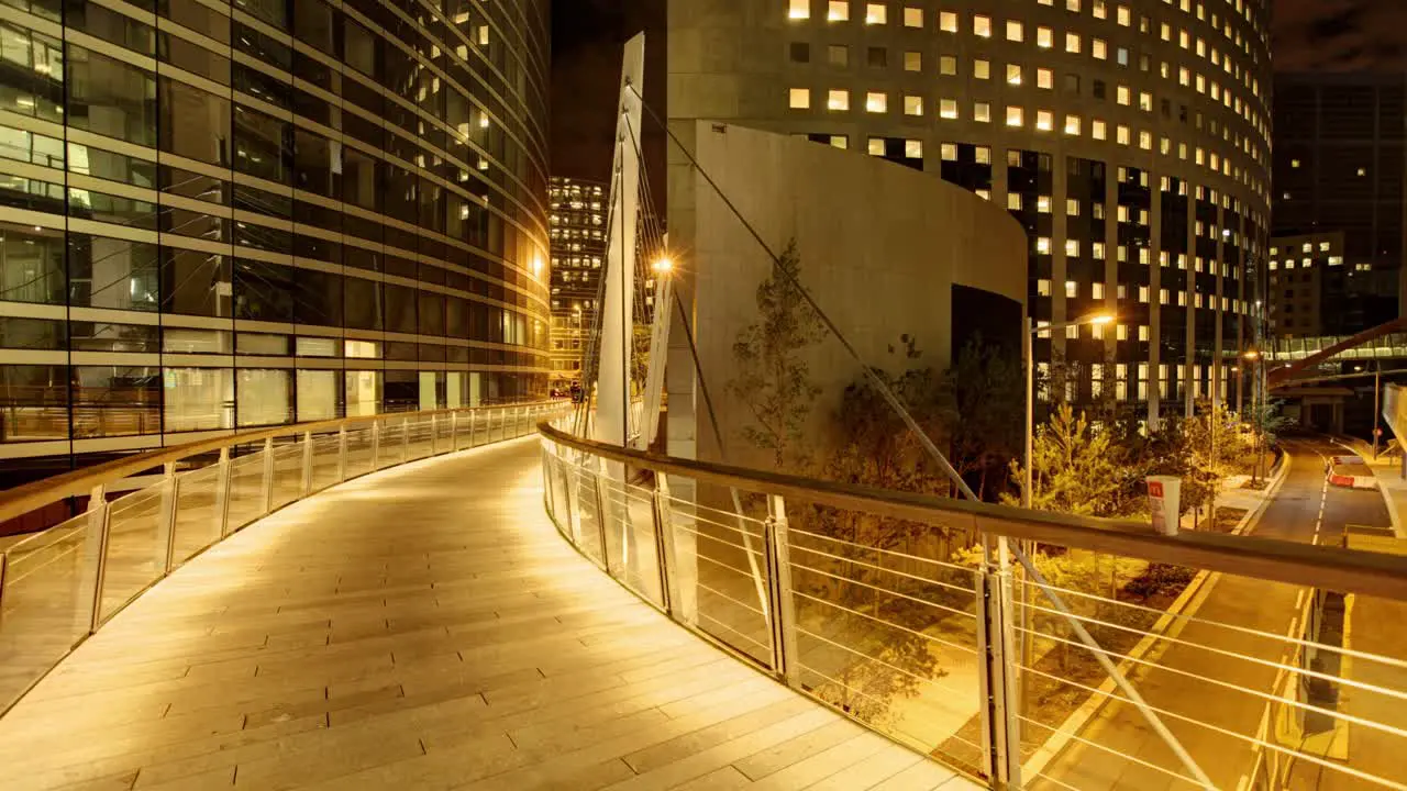 Timelapse features a warmly lit wooden pedestrian bridge with metal railing at night set against a backdrop of glowing modern buildings with curved architecture