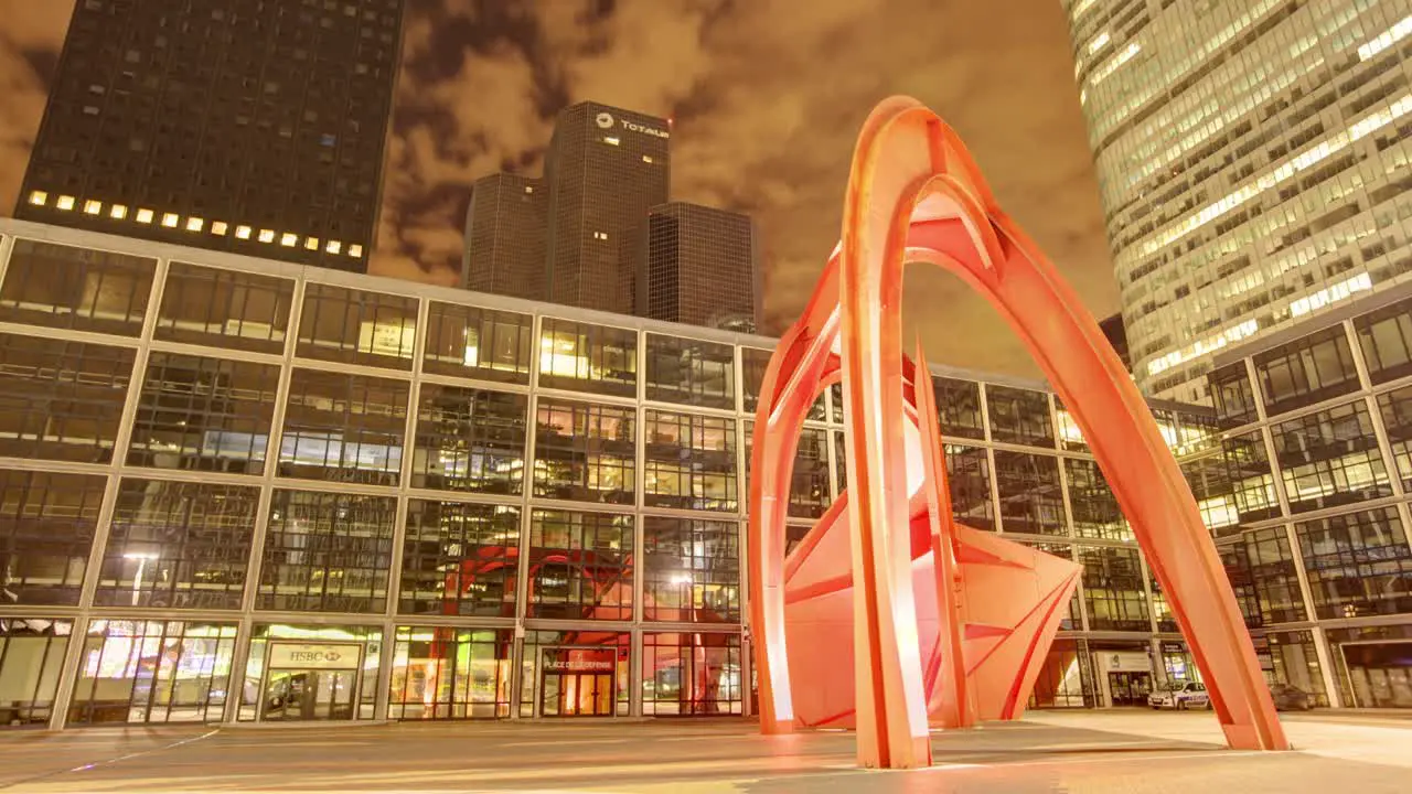 Timelapse captures an imposing large twisted orange sculpture situated against a modern grid-patterned building under a dark cloudy sky