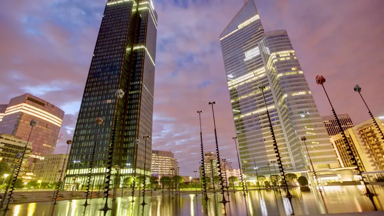 Timelapse shot captures a dreamy city skyline at night from a low angle tall modern buildings and street lamps bask in the reflection of city lights against an orange clouded sky