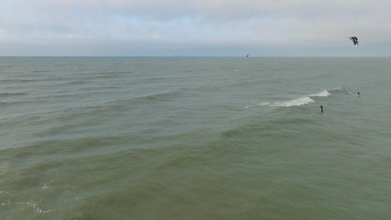 Establishing aerial view of a group of people engaged in kitesurfing overcast winter day high waves extreme sport Baltic Sea Karosta beach  distant wide drone shot moving forward