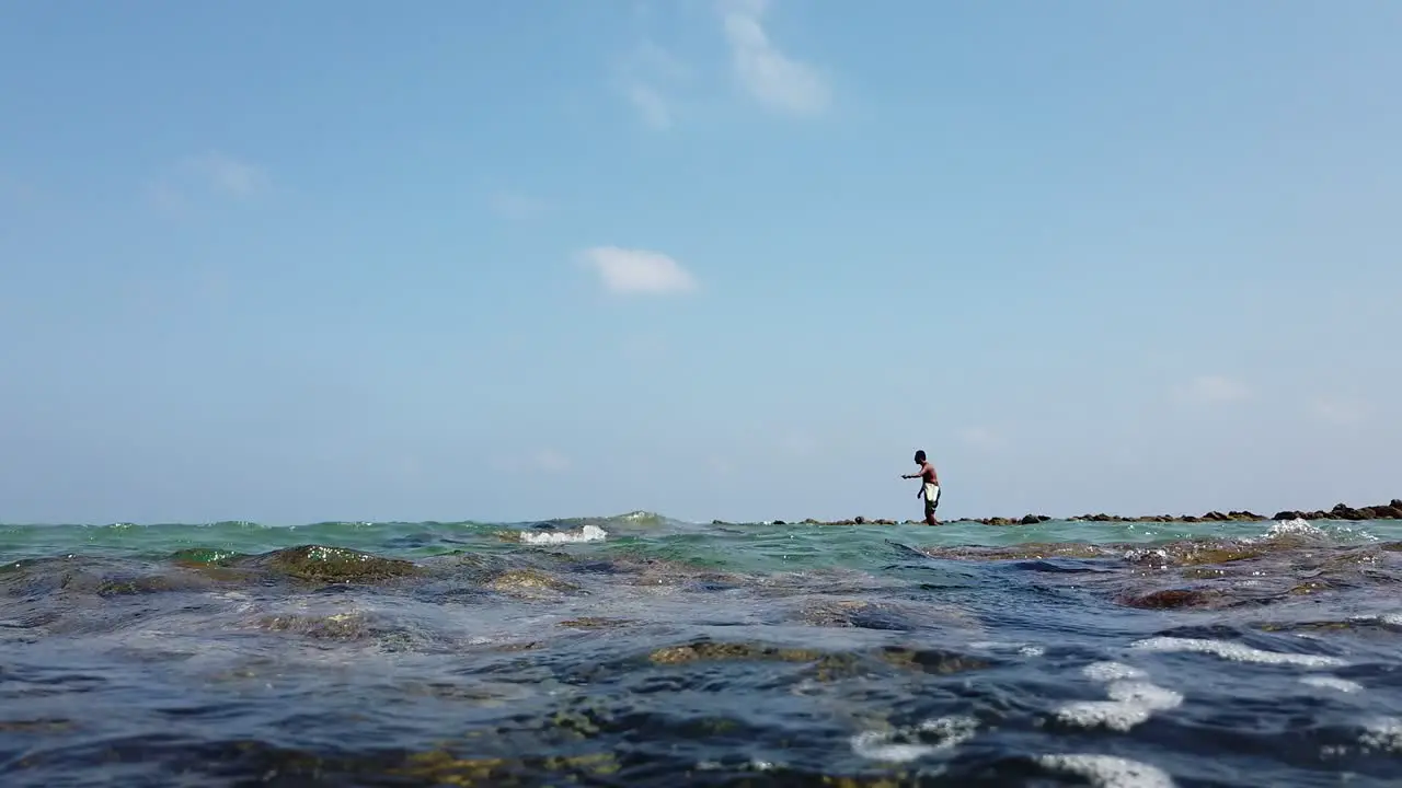 A fisherman stands on the rocks on a sunny hot day at high tide and casts a hook and line with bait again and again to catch fish to eat on the andaman islands