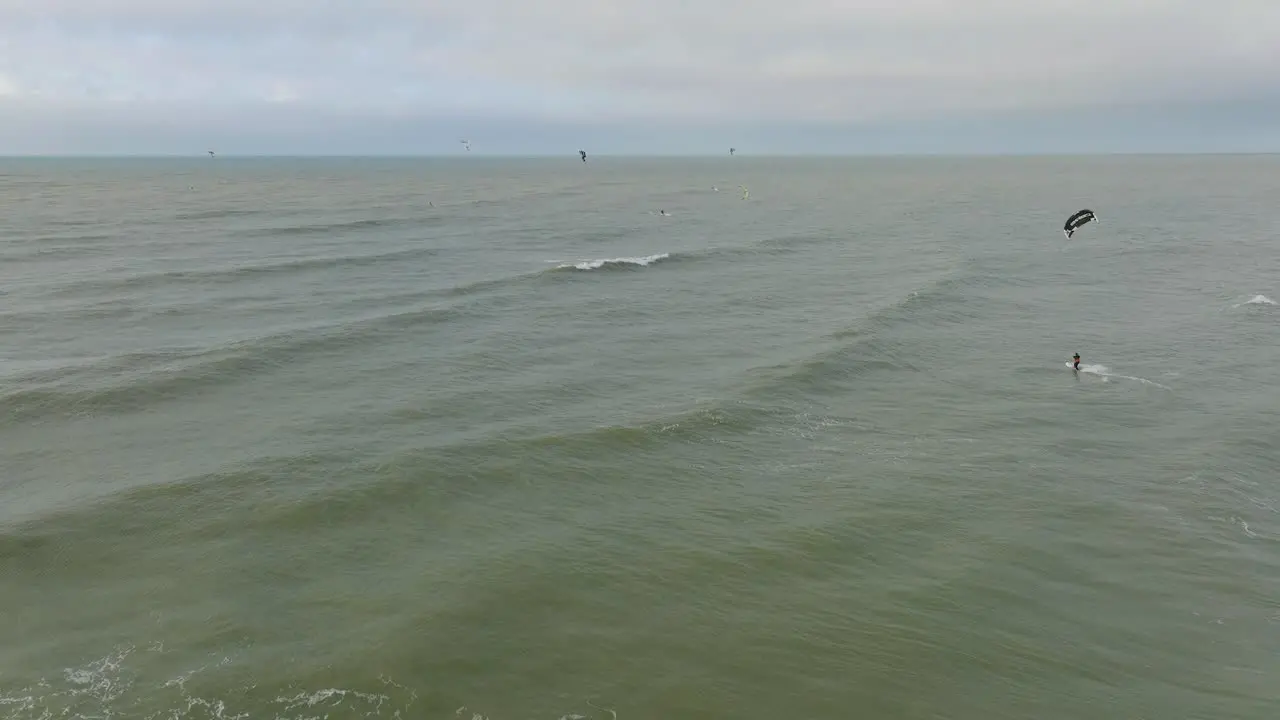 Establishing aerial view of a group of people engaged in kitesurfing overcast winter day high waves extreme sport Baltic Sea Karosta beach  wide drone shot moving forward