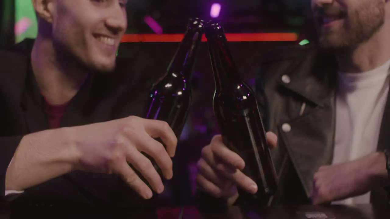 Close Up Of Two Male Friends Toasting With Beer Bottle And Talking Together While Sitting At Bar Counter