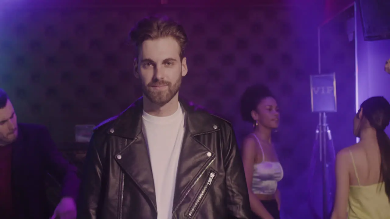 Portrait Of Handsome Bearded Man In Leather Jacket Smiling At Camera At Disco While His Friends Dancing Behind Him