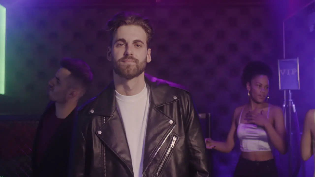 Portrait Of Handsome Bearded Man In Leather Jacket Smiling At Camera At Disco While His Friends Dancing Behind Him 1