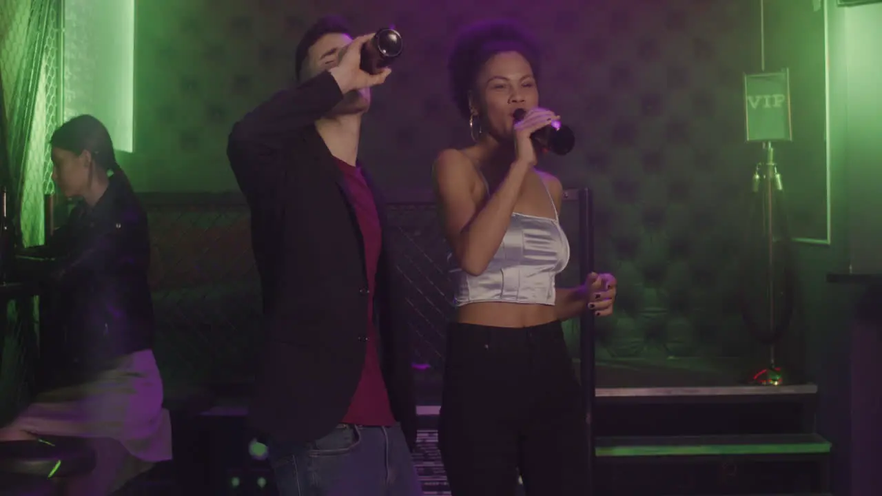 Happy Multiethnic Couple Drinking Beer And Dancing At Disco While A Serious Girl Sitting On A Stool And Using Mobile Phone