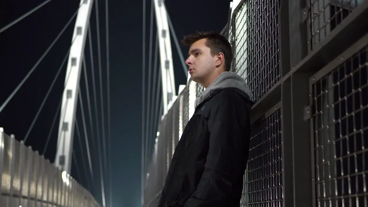 Young white male teen standing on a bridge at night looking around