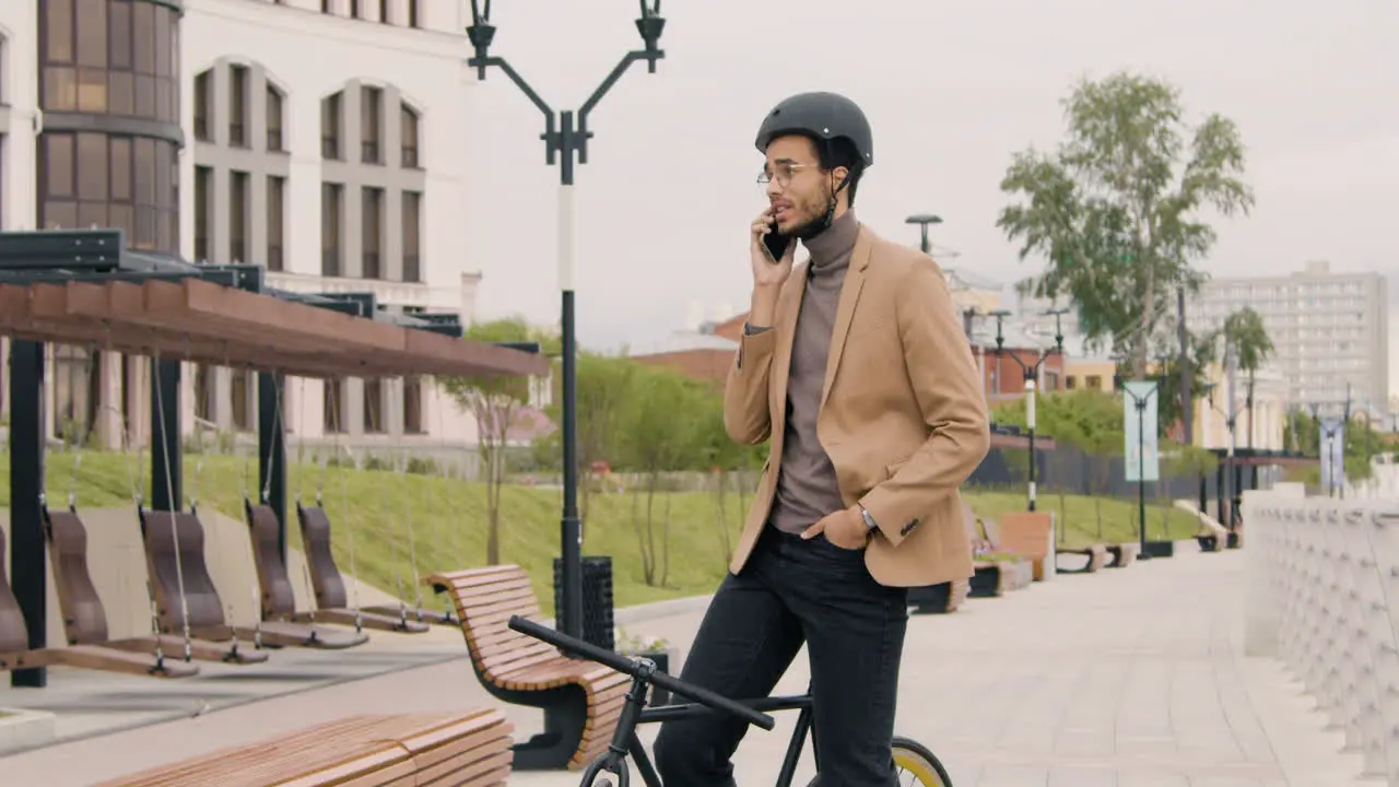 Young American Man In Formal Clothes And Helmet Having A Call On Mobile Phone While Sitting On Bike In The City