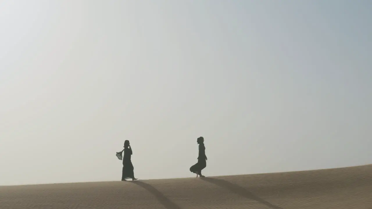 Extreme Long Shot Of Two Muslim Women Wearing Traditional Black Dress And Hijab Walking In A Windy Desert 1