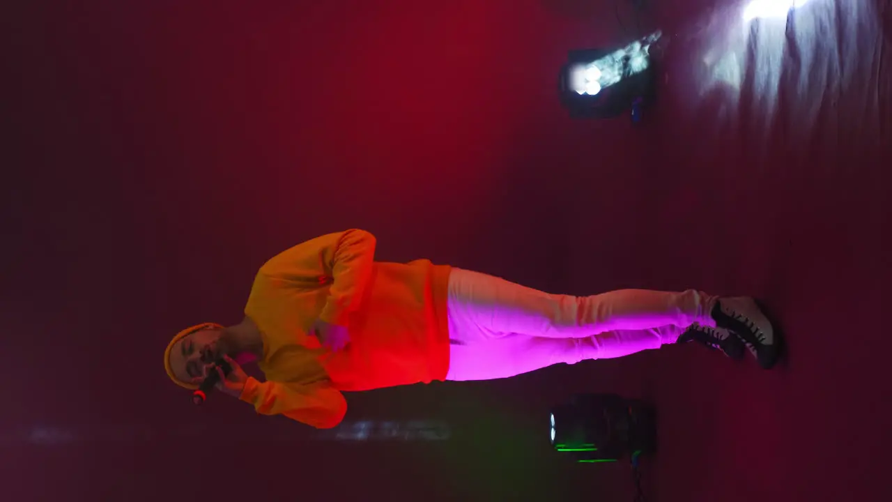 Vertical video Funny male rap artist sings and dances into a microphone on a stand in the neon light of lamps and in the Studio on a colorful bright background