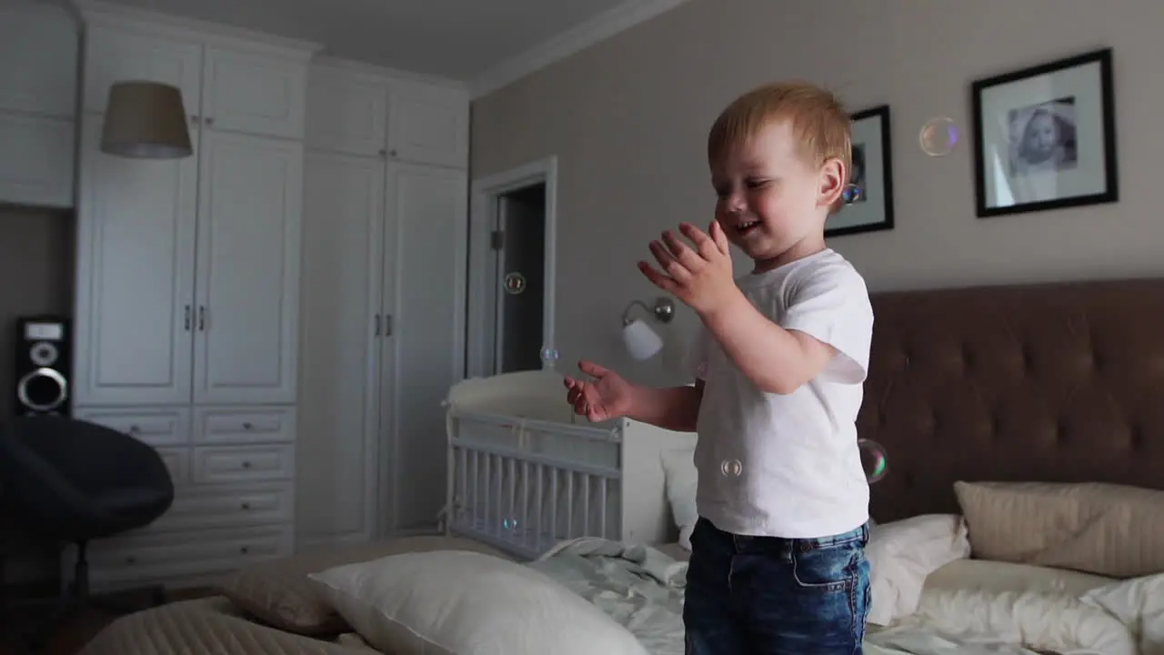 A boy in a white T-shirt jumps on the bed and laughs at the sight of soap bubbles in the parents' bedroom in slow motion