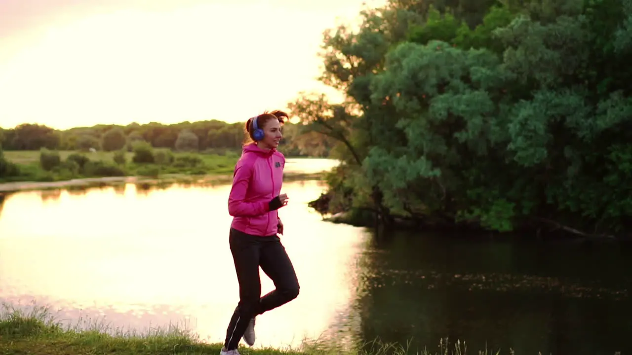 Brunette with long hair in headphones runs along the river in the Park in the morning at sunrise in the summer in a pink jacket and black pants