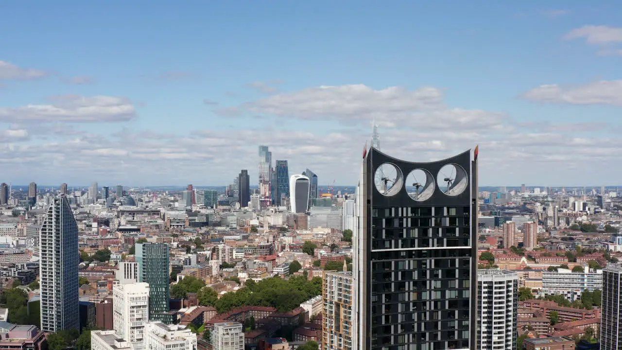 Forwards fly around modern skyscraper with wind turbines in top part of construction Revealing panoramic view of city with group of tall office buildings London UK