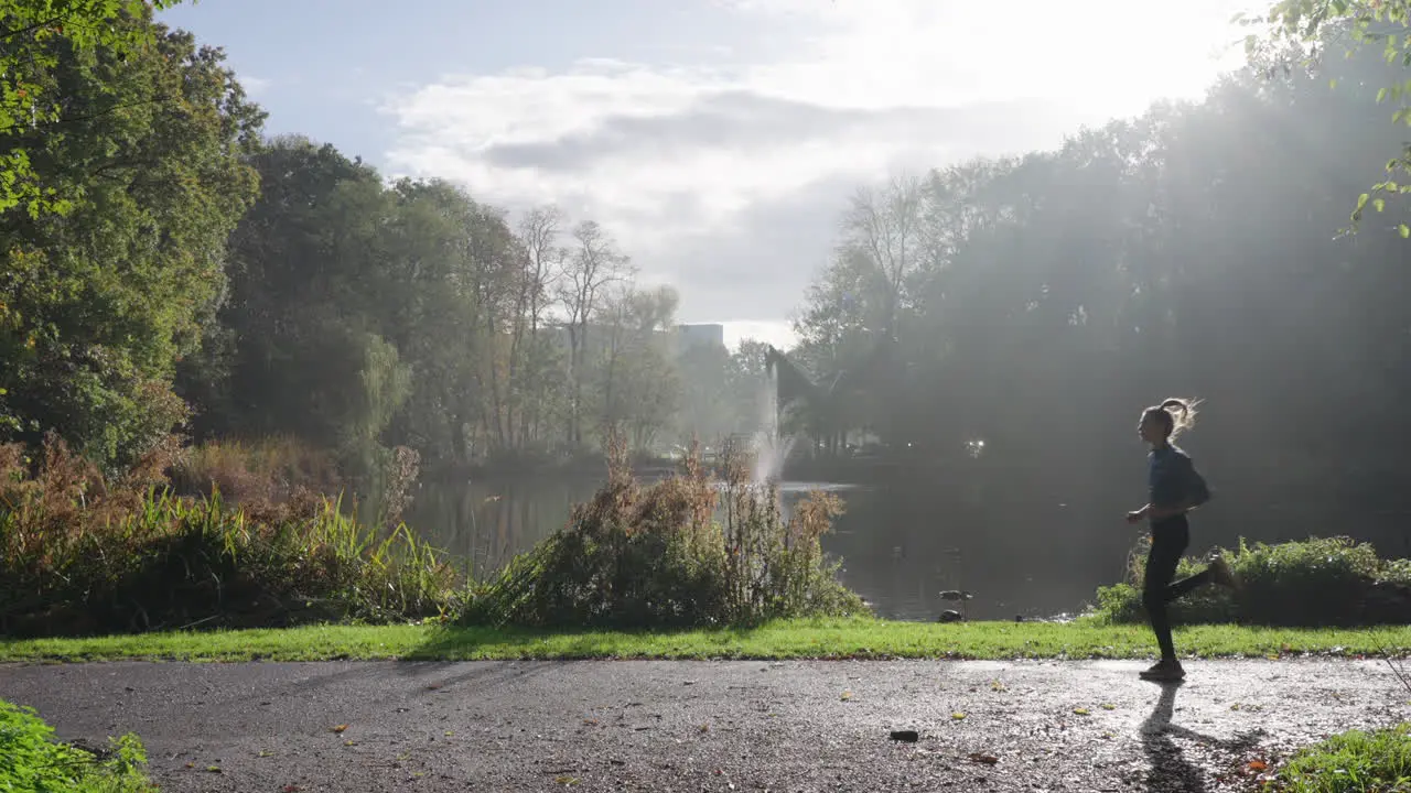 This stock footage captures a woman running from right to left in a sunny park with a pond in the background outdoor exercise amidst beautiful natural surroundings