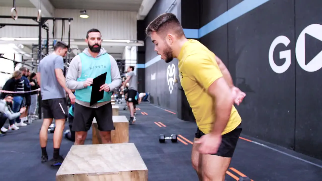 a young Caucasian man performing jump boxes in a crossfit gym under the supervision of an evaluator during a crossfit competition