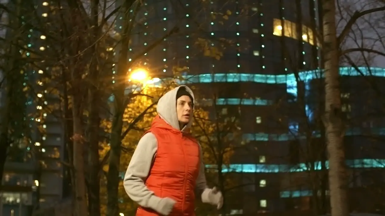 Camera Focuses On A Woman In Sportswear And Hood Running In The City At Night In Winter