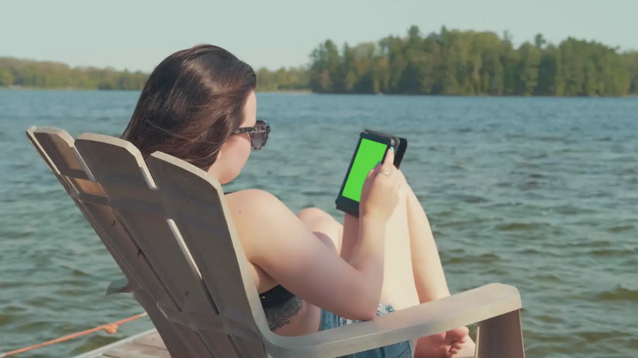 Camera pans over a woman relaxing on a dock next to a lake in the summer season with green screen for chroma key