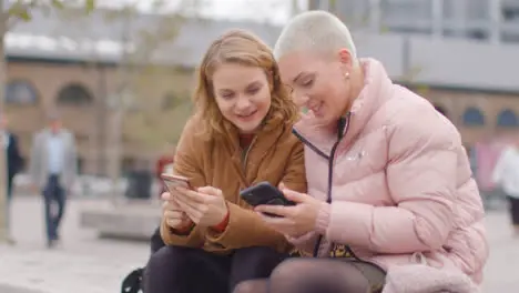 Tracking Shot of Two Friends Sitting Outside On Their Phones