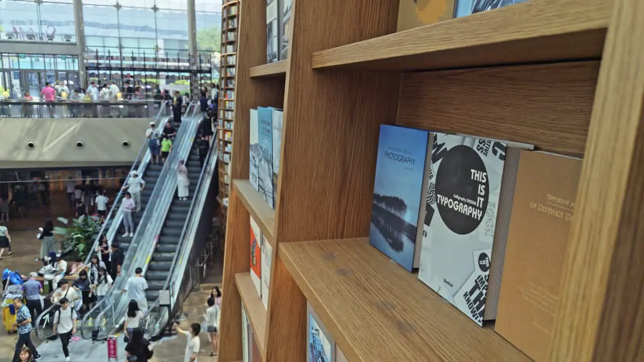 Focus on books in shelf as crowds come down the escalator