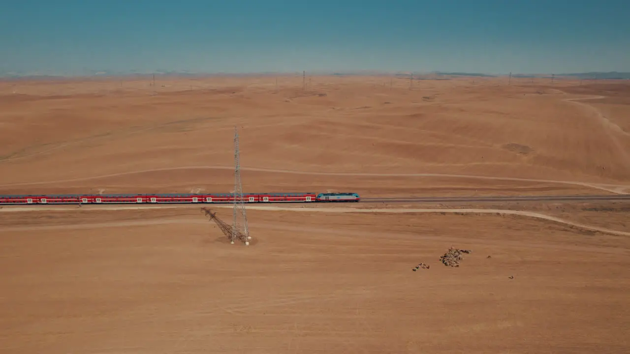 Aerial shot of a red passenger train in a remote and desert area near large electricity poles dry land without crops on the ground there is a female shepherd with sheeps