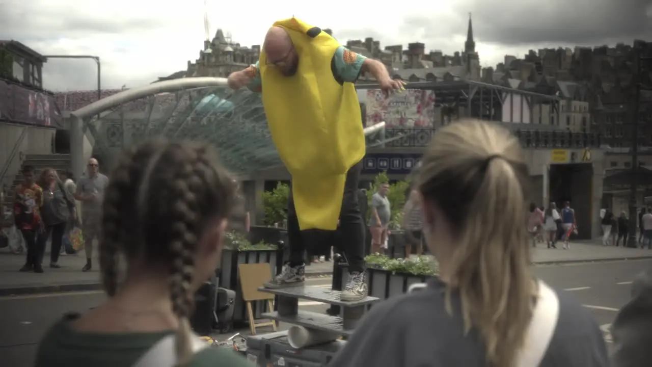 Witness an extraordinary man performing a captivating balancing act in the vibrant city of Edinburgh captivating a crowd as two girls watch in the foreground