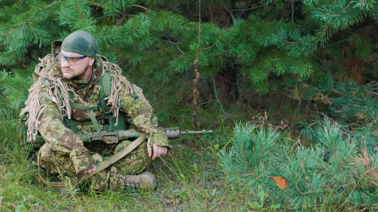 The Young Man In Uniform With A Weapon In His Hands He Is Sitting In The Forest Resting Looking Arou