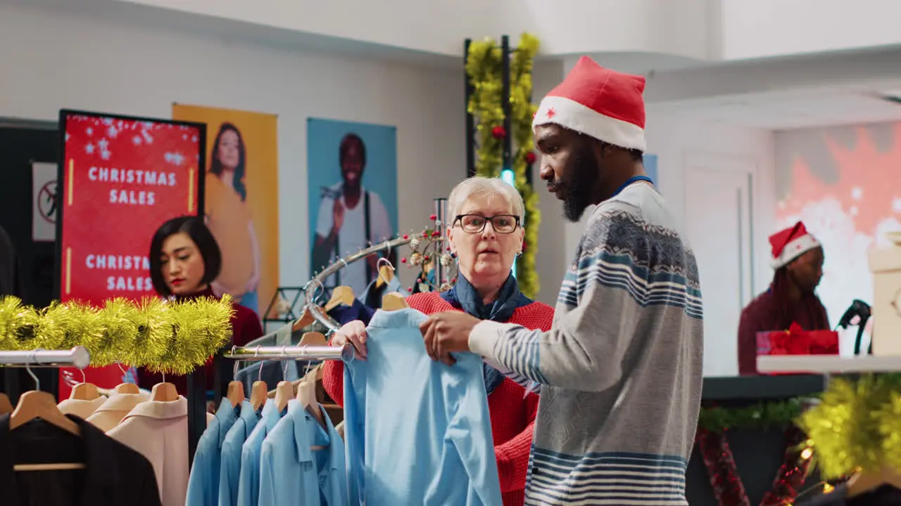 African american worker wearing Santa hat helping woman browsing for clothes in festive Christmas decorated clothing store Employee assisting elderly customer in festive adorn fashion shop