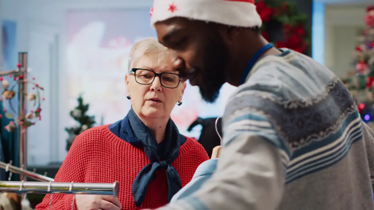 African american employee wearing Santa hat in festive Christmas decorated clothing store offering shopping advice to indecisive senior customer during xmas holiday season