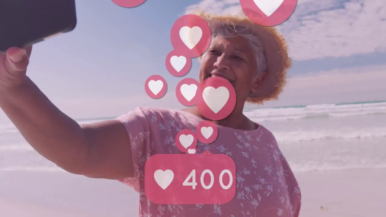 Animation of hearts over happy senior african american woman taking selfie on sunny beach