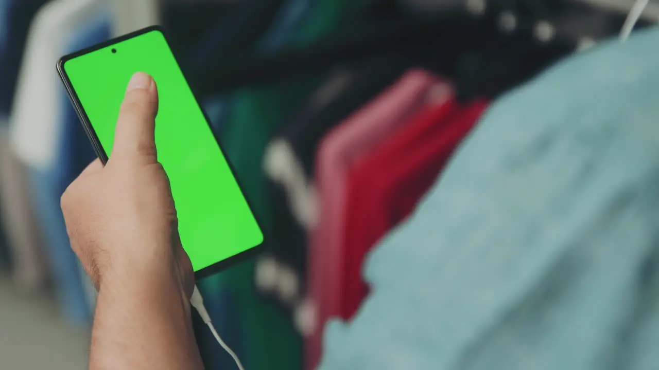 Man in Shirt Using Mobile Phone with Green Screen Scrolling through the Display while Watching and Listening through the Headphones in a Clothes Store