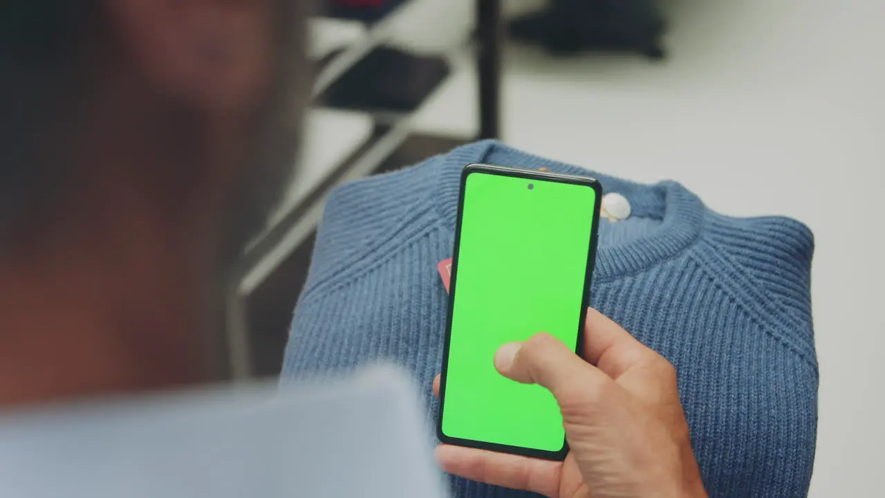 Elegant man dressed in light blue shirt scrolling through a green screen phone while holding a blue sweater in a clothes store