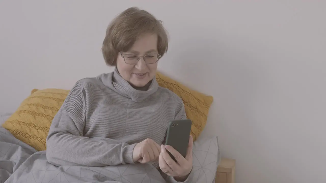 Senior Woman With Eyeglasses Sitting On Bed Making A Video Call On A Smartphone