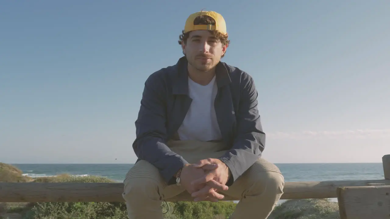 Handsome Man In Cap Sitting On Boardwalk Looking At Camera