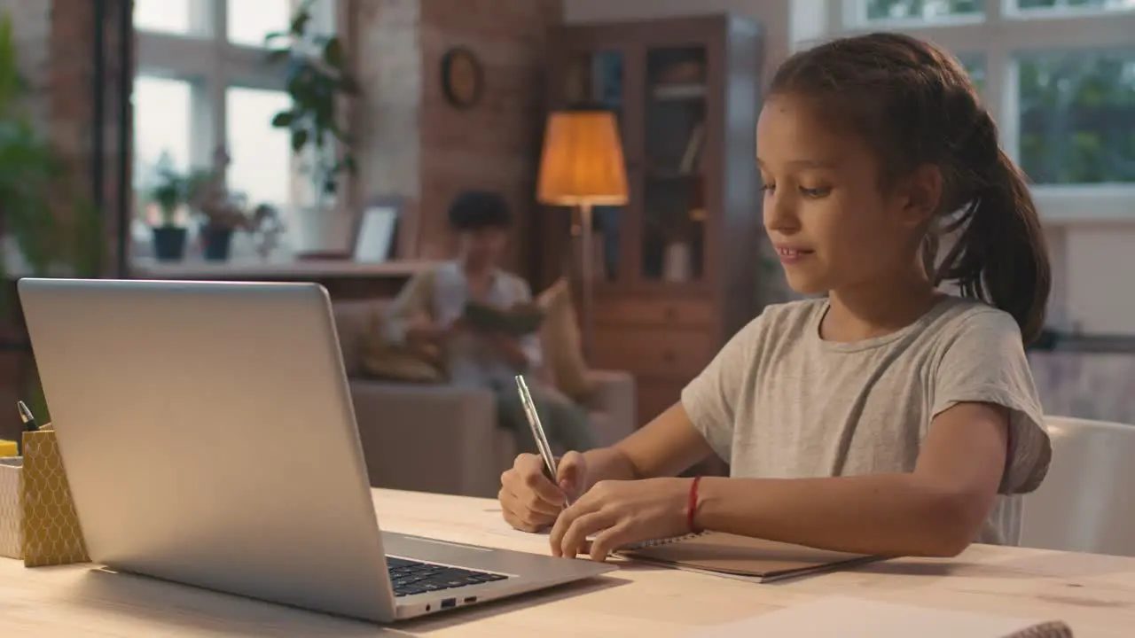 Brunette Girl Sitting At A Desk In Front Of The Computer On A Video Call While Taking Notes Behind Her Mother Sits In An Armchair