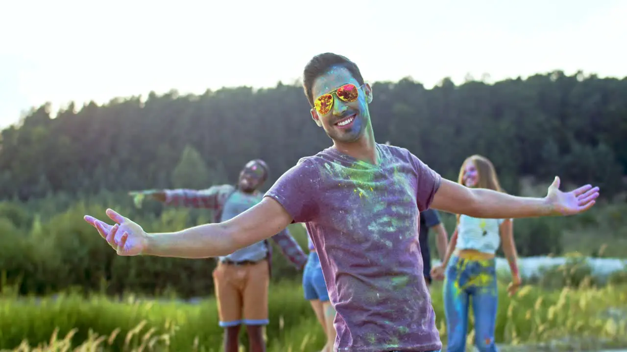 Portrait Of The Young Handsome Man In Sunglasses And All In Colorful Paints Spots Smiling To The Camera And Posing Outdoors While Having Festive Holi Day With Multiethnic Friends