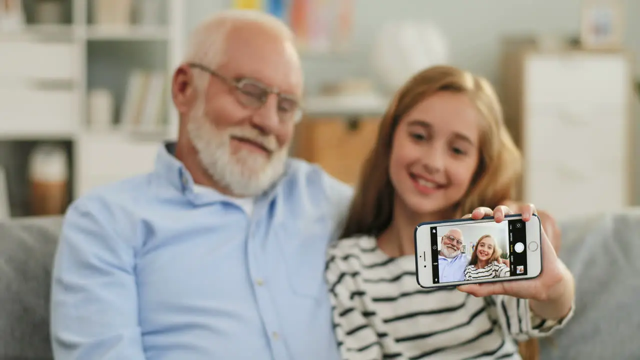 Portrait Of The Pretty Schoolgirl Sitting On The Sofa With A Grandfather In Glasses Smiling And Taking Selfie Photo On The Smartphone Camera