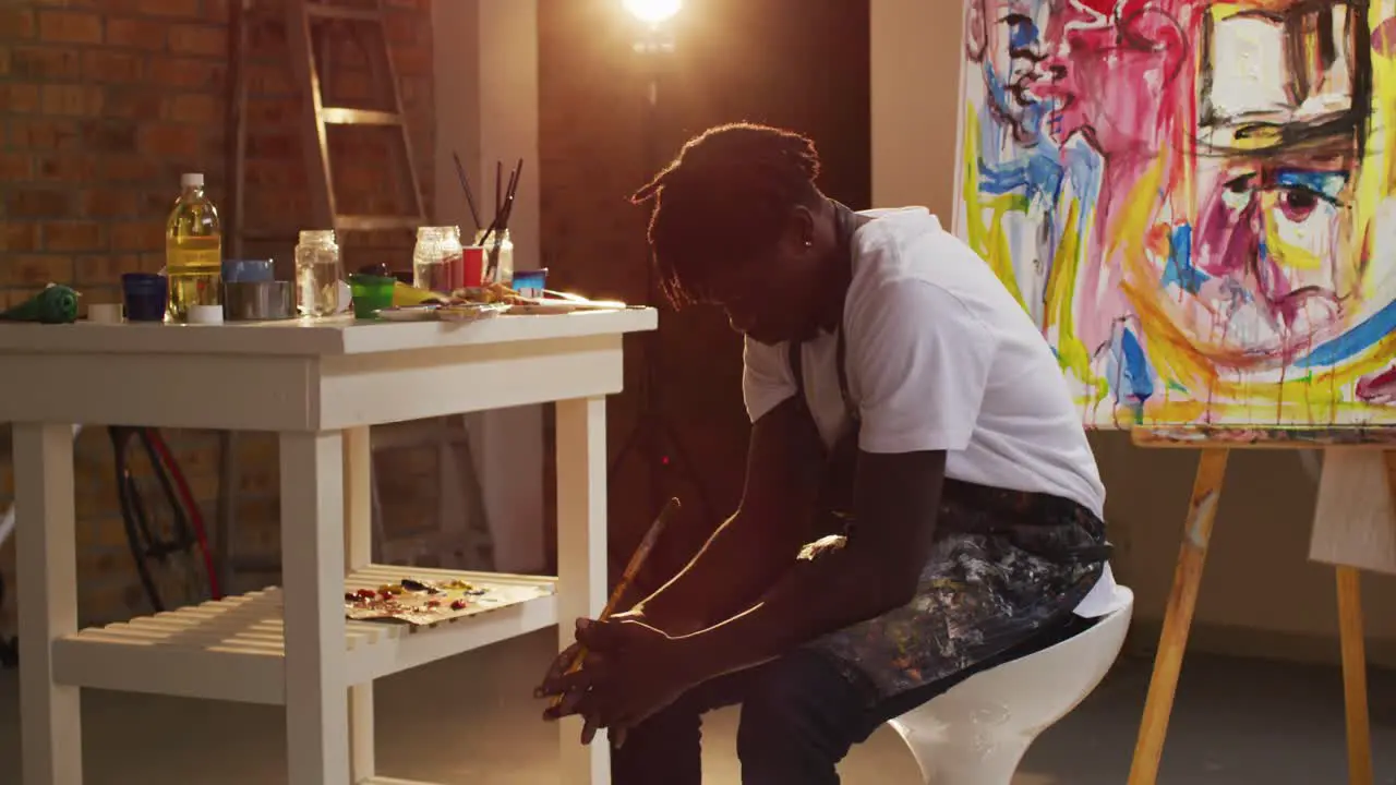 Stressed african american male artist wearing aprons sitting on a stool at art studio