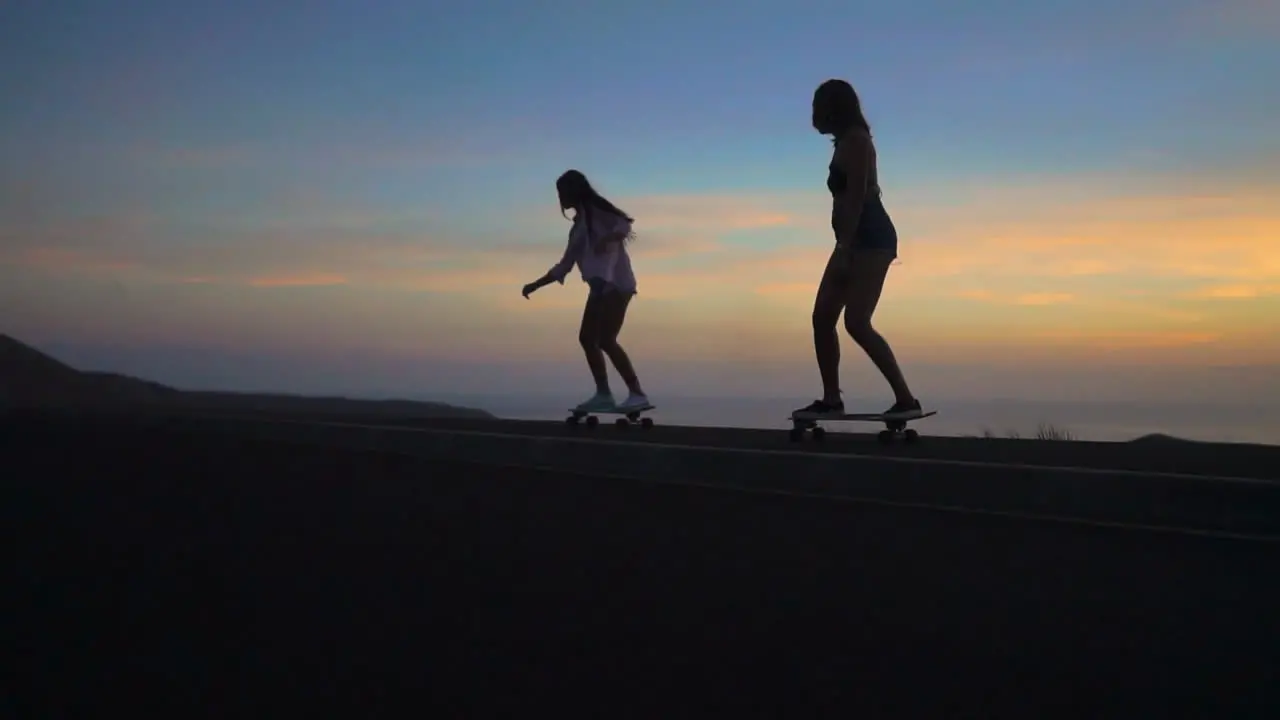 Two friends skate along a road at sunset their movements captured in slow motion against the backdrop of mountains and a breathtaking sky They're wearing shorts