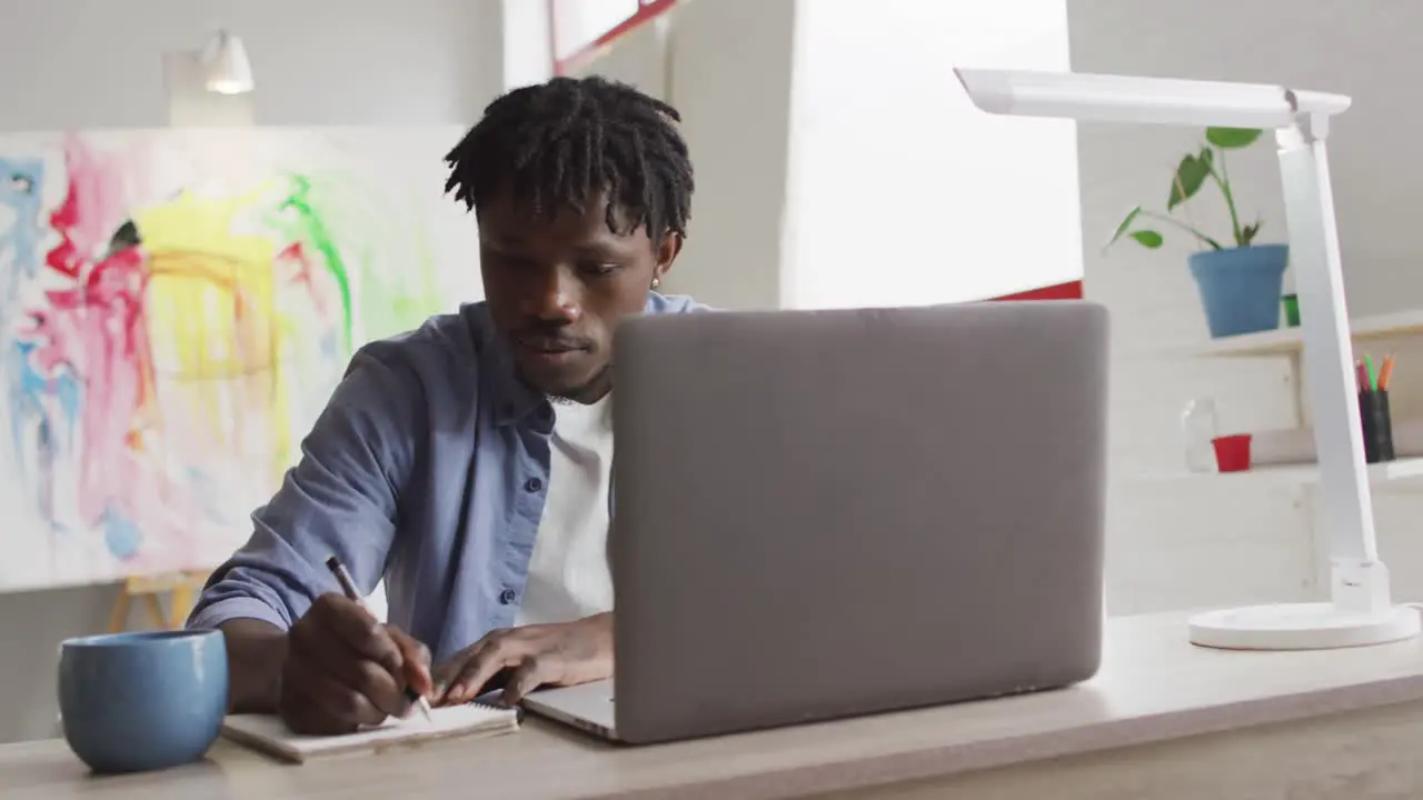 African american male artist with laptop taking notes while sitting on his desk at art studio