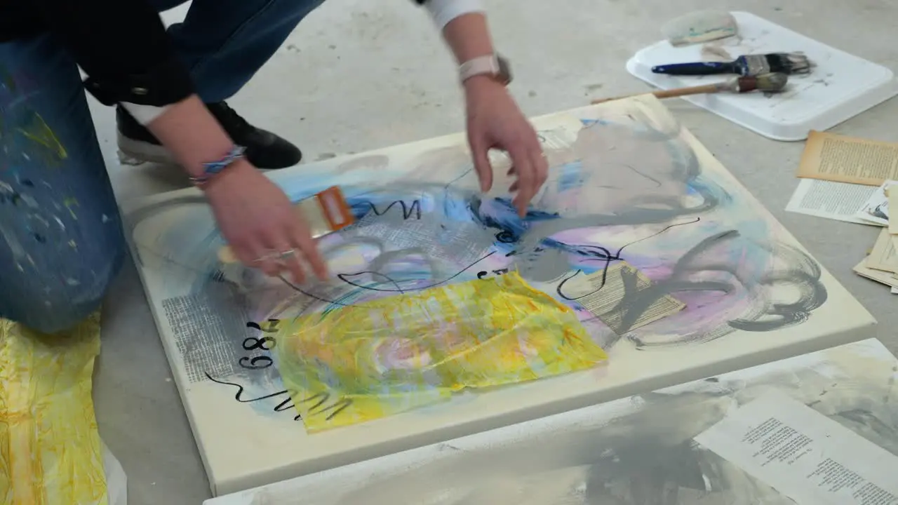 Female artist on floor of a busy home studio layering yellow paper on a canvas