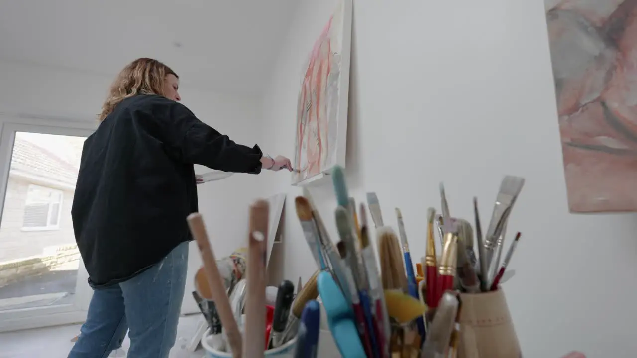 Artist stands painings canvas in home studio foregrounded by pots of brushes